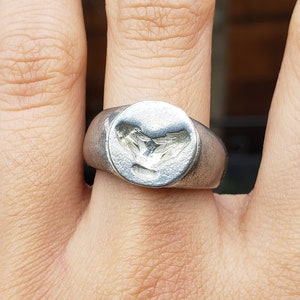 May include: A silver ring with a heart-shaped design on the top. The ring is worn on a person's finger.