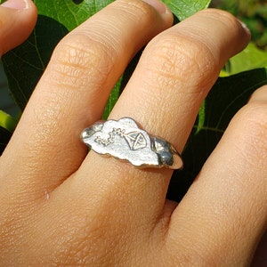 May include: A silver ring with a cloud design. The cloud has a small tree and a sun inside of it.