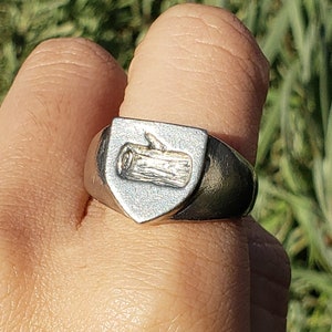 May include: A silver ring with a shield-shaped design featuring a stylized log inside. The ring is worn on a person's finger.