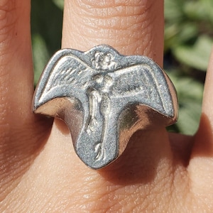 Puede incluir: Un anillo de plata con un diseño detallado de una figura alada. El anillo se lleva en el dedo de una persona.