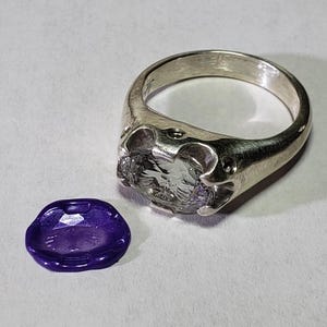 May include: A silver-toned metal ring featuring a wide, textured band and a prong setting that holds a clear, faceted oval gemstone. Beside the ring lies a small, flat, purple wax seal with a subtle embossed design on its surface.