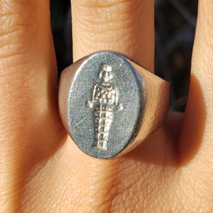 Puede incluir: Anillo de sello plateado con una cara ovalada. El anillo presenta una figura en relieve detallada. El anillo se lleva en un dedo, mostrando el diseño y la artesanía. El metal del anillo tiene un aspecto ligeramente envejecido.