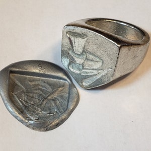 Puede incluir: Un anillo de plata con un diseño detallado en la parte superior. El anillo se muestra junto a un sello de plata con un diseño similar.