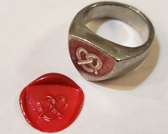 May include: A silver ring with a pretzel design on the top. The ring is next to a red wax seal with a pretzel design.