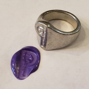 May include: A silver ring with a raised letter "P" and a small diamond. A purple wax seal with the same letter "P" is next to the ring.
