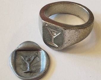 Puede incluir: Un anillo de sello de plata con un diseño de copa de martini. El anillo se muestra junto a un sello de cera con el mismo diseño.