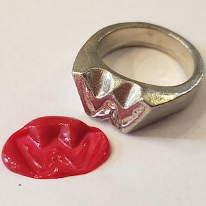 Puede incluir: Un anillo de plata con una letra "W" en relieve en la cara. El anillo se muestra junto a un sello de cera roja con el mismo diseño de la letra "W".