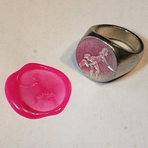 May include: A silver signet ring with a pink oval face featuring a relief of three figures. A bright pink wax seal with a similar design is next to the ring. The ring and seal are on a white surface.