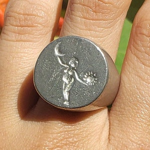 May include: A silver signet ring with a raised design of a figure holding a crescent moon and a flower.
