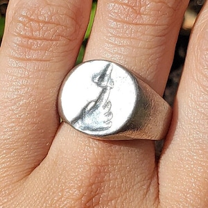 May include: A silver ring with a raised design of a hand giving the middle finger. The ring is worn on the middle finger of a person's hand.