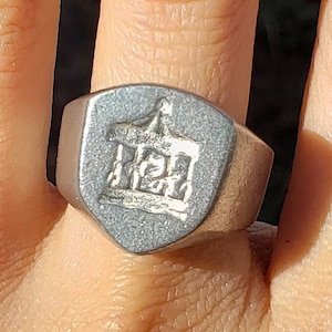 Op de afbeelding: Een zilverkleurige zegelring met een schildvormige voorkant. De voorkant heeft een gedetailleerd, verhoogd ontwerp van een structuur met een toren en andere architectonische elementen. De ring wordt aan een vinger gedragen.