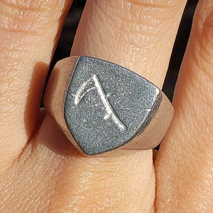 May include: A silver signet ring with a shield-shaped design featuring a stylized symbol. The ring is worn on a person's finger.