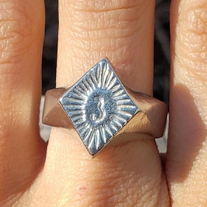 May include: A silver ring with a diamond-shaped face. The face features a sunburst design around the number "3". The ring is worn on a finger, showcasing its geometric design and metallic sheen. The ring's design is simple yet eye-catching.