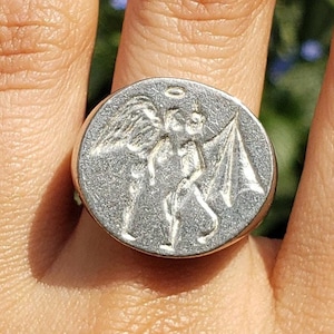 May include: A silver ring featuring a detailed design of an angel and a devil on the face of the ring.