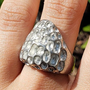 May include: A silver ring with a textured, organic-shaped design. The ring is worn on a person's finger.
