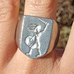 以下が含まれることがあります： 剣を持った女性が描かれた盾の形をしたデザインの銀の指輪。女性は戦士のポーズで描かれており、盾は縁が高くなっている。