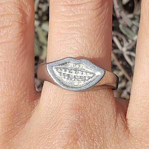 May include: A silver ring with a raised design of a mouth with teeth. The ring is worn on a person's finger.