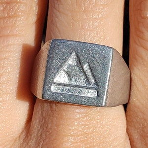 May include: A silver signet ring with a square face featuring a raised, stylized mountain design.