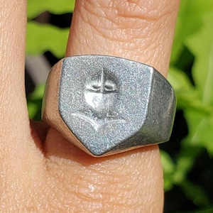May include: A silver-colored signet ring with a shield-shaped face. The face features an embossed design of a cross above an anchor. The ring is worn on a finger, with green foliage in the background.