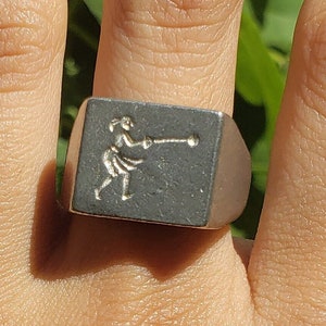 Puede incluir: Un anillo de plata con una cara cuadrada que presenta un diseño en relieve de una persona lanzando una pelota.