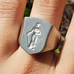 May include: A silver-colored signet ring with a shield-shaped face. The face features a detailed relief of a person holding a staff. The ring is worn on a finger, with a blurred green background.