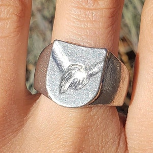 May include: A silver ring with a raised design of two hands clasped together. The ring is worn on a person's finger.
