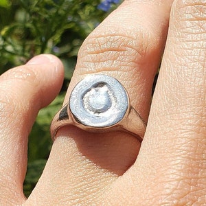 May include: A silver ring with a circular face featuring a raised, stylized design. The ring is shown on a hand, with a blurred background of green foliage and small blue flowers.