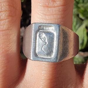 May include: A silver signet ring with a rectangular design featuring a stylized leaf inside. The ring is worn on a person's finger.