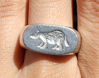 May include: Silver ring with a rectangular face. The ring has a detailed engraving of a badger. The ring is worn on a finger, showcasing the badger design. The ring's metal has a matte finish.