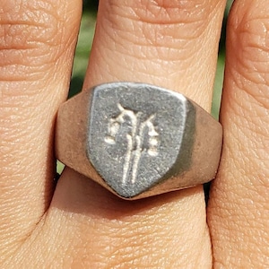 May include: A silver signet ring with a shield-shaped design featuring a stylised tree with three branches. The ring is worn on a person's finger.