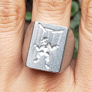 May include: A silver ring with a square face featuring a raised design of a puppet with strings attached. The puppet is in a stylized pose with its arms outstretched.