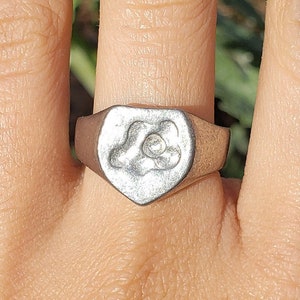 May include: A silver signet ring with a heart-shaped design featuring a stylized cloud with a small circle in the center.