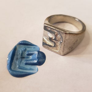 May include: A silver signet ring with a raised design of a stylised figure. The ring is shown next to a blue wax seal impression of the same design.