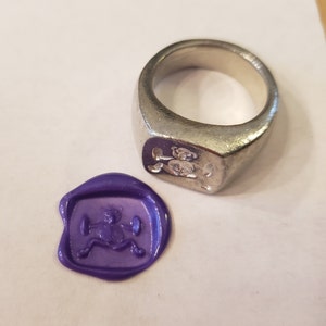 May include: A silver signet ring with a design of a bear holding a dumbbell. The ring is shown next to a purple wax seal with the same bear design.