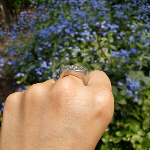 May include: A silver signet ring with an engraved design. The ring is worn on a person's finger.