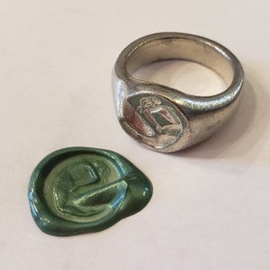 May include: A silver ring with a green wax seal impression. The ring has a raised design on the top. The wax seal is a green circle with a raised design in the center.