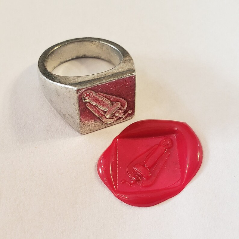 May include: A silver signet ring with a raised design of a seated figure. The ring is shown next to a red wax seal with the same design.
