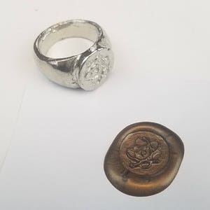 May include: A silver ring with a detailed design on the top. The ring is next to a wax seal with the same design.