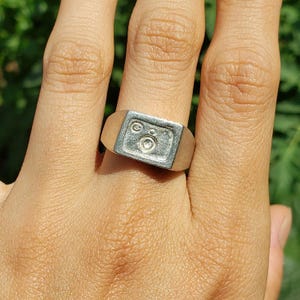Puede incluir: Un anillo de plata con un diseño cuadrado que presenta un círculo con un círculo más pequeño en su interior. El anillo se lleva en un dedo.
