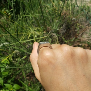 May include: A silver metal ring with a square shape. The ring is worn on a person's finger.