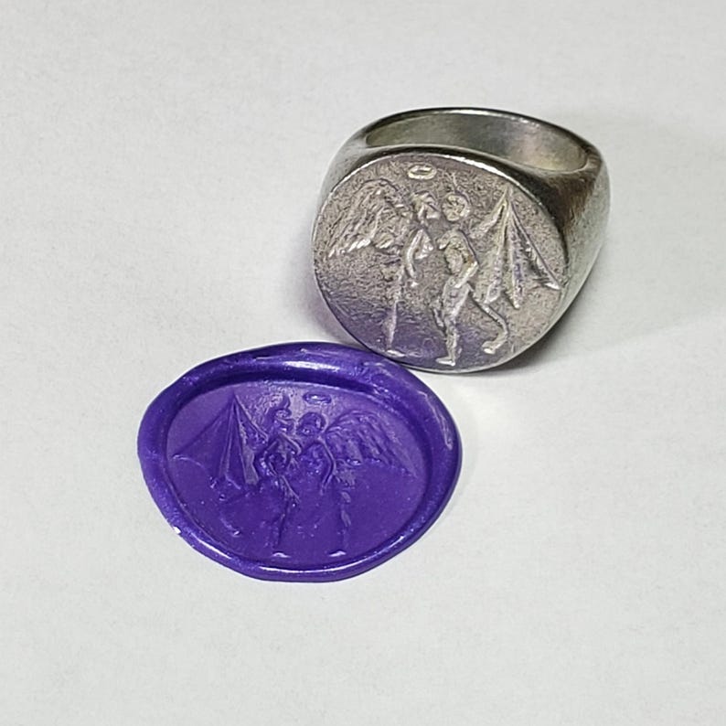 May include: A silver signet ring with a detailed design of an angel with wings and a halo. The ring is shown next to a purple wax seal with the same design.