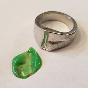 May include: A silver ring with a wide, flat band and a rounded, slightly pointed top. A small green blob of what appears to be slime or putty is next to the ring.