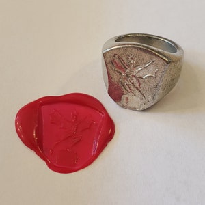 May include: A silver signet ring with a raised design of a winged creature. The ring is shown next to a red wax seal with the same design.