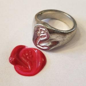 Puede incluir: Un anillo de sello de plata con un diseño detallado en la cara. El anillo se muestra junto a un sello de cera roja con el mismo diseño.