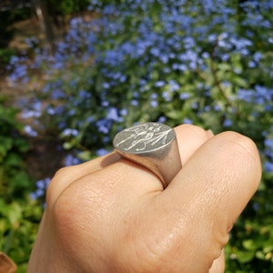 May include: A silver ring with a detailed design of two figures, possibly angels, with wings and a spear. The ring is worn on a person's finger.