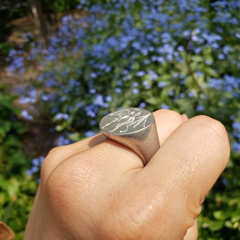 May include: A silver ring with a detailed design of two figures, possibly angels, with wings and a spear. The ring is worn on a person's finger.