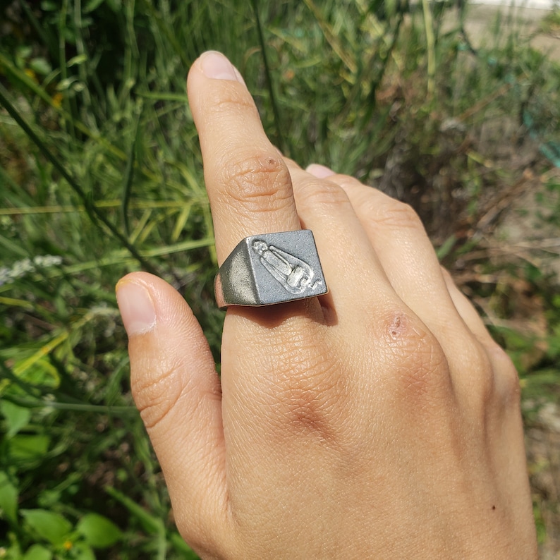 May include: A silver ring with a square face featuring a raised, detailed design of a person in a praying position.
