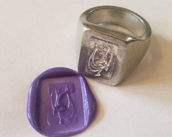 May include: A silver-toned metal signet ring featuring a rectangular face with an engraved figure of a person dancing. Beside the ring is a purple wax seal impression that mirrors the same engraved dancing figure design in a rectangular shape.
