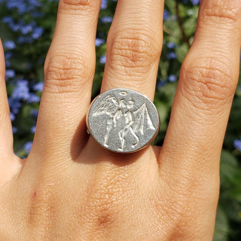 May include: A silver ring with a detailed design of an angel and a devil on the face of the ring.