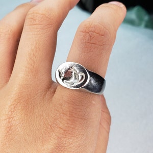 Puede incluir: Un anillo de plata con un diseño en relieve de una tetera en la parte superior. El anillo se lleva en el dedo de una persona.